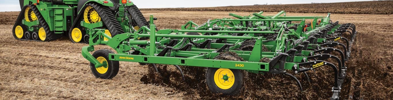 Green John Deere tractor with yellow wheels plowing a flat field under an overcast sky.
