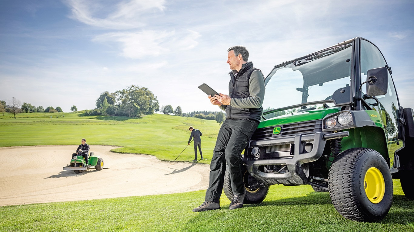 Person leaning on to gator holding a tablet looking at the workers raking the golf turf
