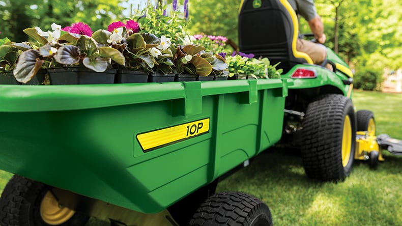 A John Deere ride on lawn mower pulling a cart full of flowers.