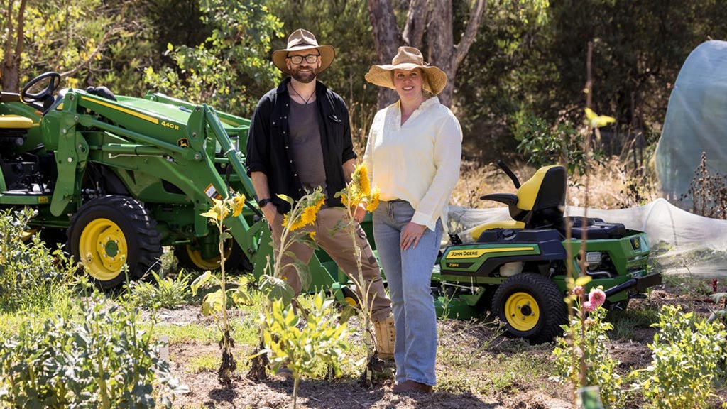 Two people in muddy farmyard with tractor and greenhouse in background.