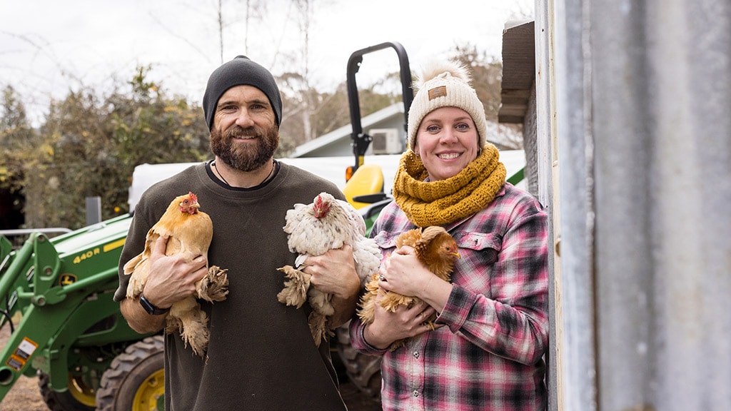 Two people in muddy garden with film crew, greenhouse and trees in background.