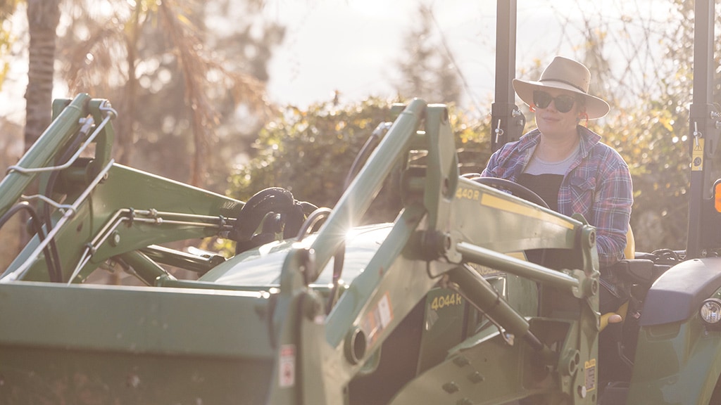 Two people in hats in garden with green tractor, lawnmower, and surrounding plants.