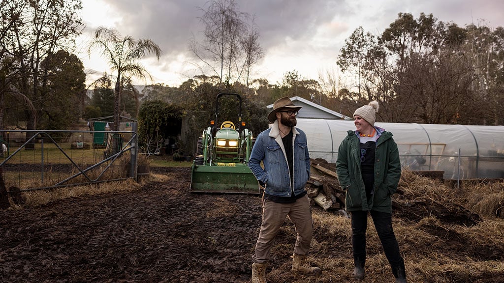 Two people holding chickens near a green tractor, one in black beanie, one in yellow scarf.