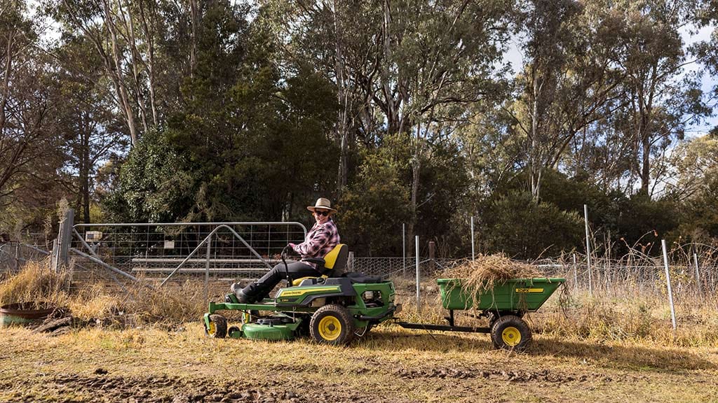 Person on green John Deere lawn tractor pulling hay-filled trailer, wearing hat and plaid shirt.
