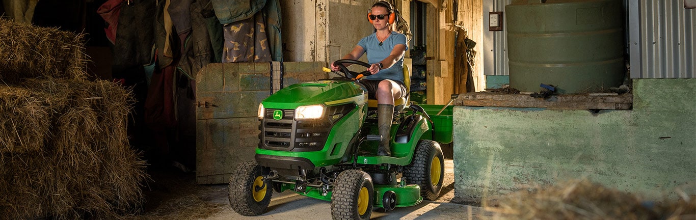 Person with ear protection drives green John Deere lawn tractor in barn with hay bales.