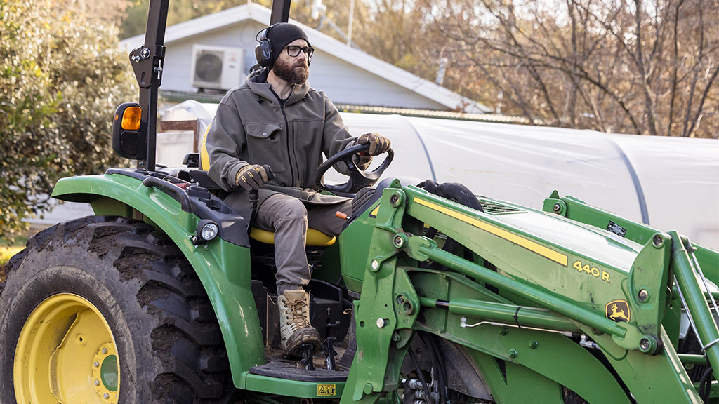 Person with headphones and glasses drives green John Deere 440R tractor outdoors.