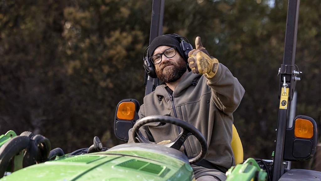 Person in beanie and gloves gives thumbs-up while sitting on green tractor.