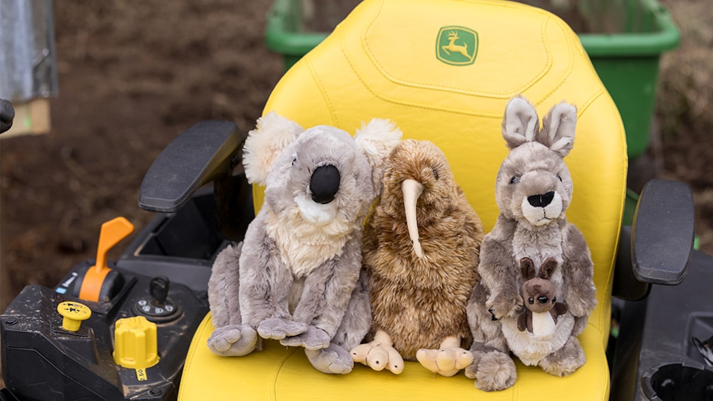 Three stuffed animals—koala, kiwi, and kangaroo—sit on yellow seat of John Deere tractor.