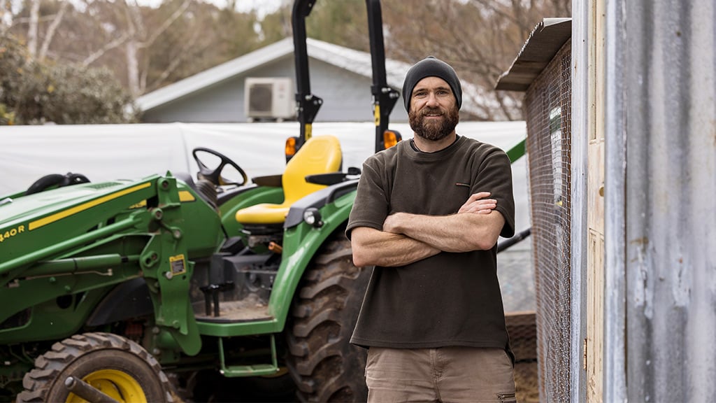 Two people holding chickens stand near green tractor and chicken coop in outdoor setting.