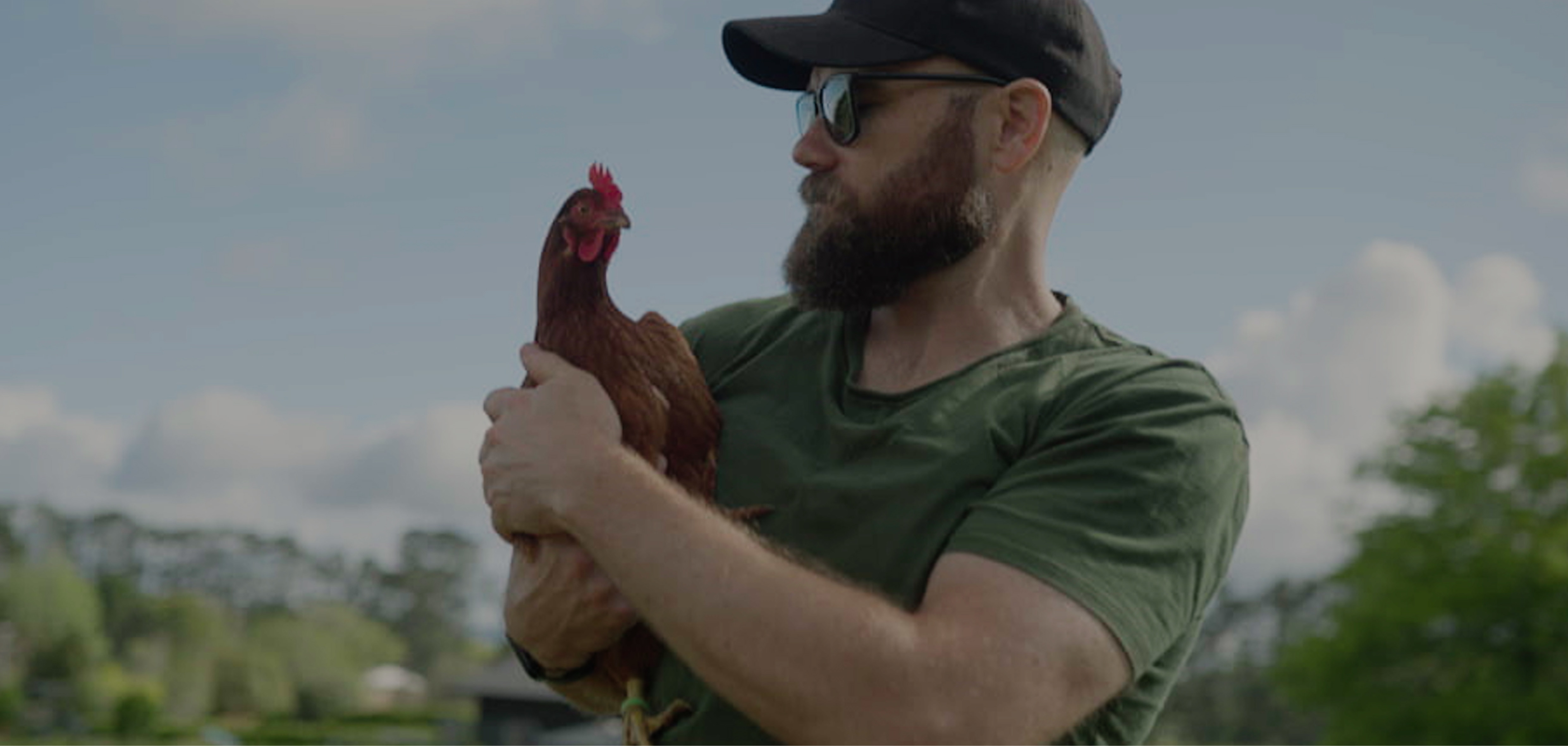 Man wearing sunglasses and a hat in agreen shirt with a beard holding a chicken with both hands