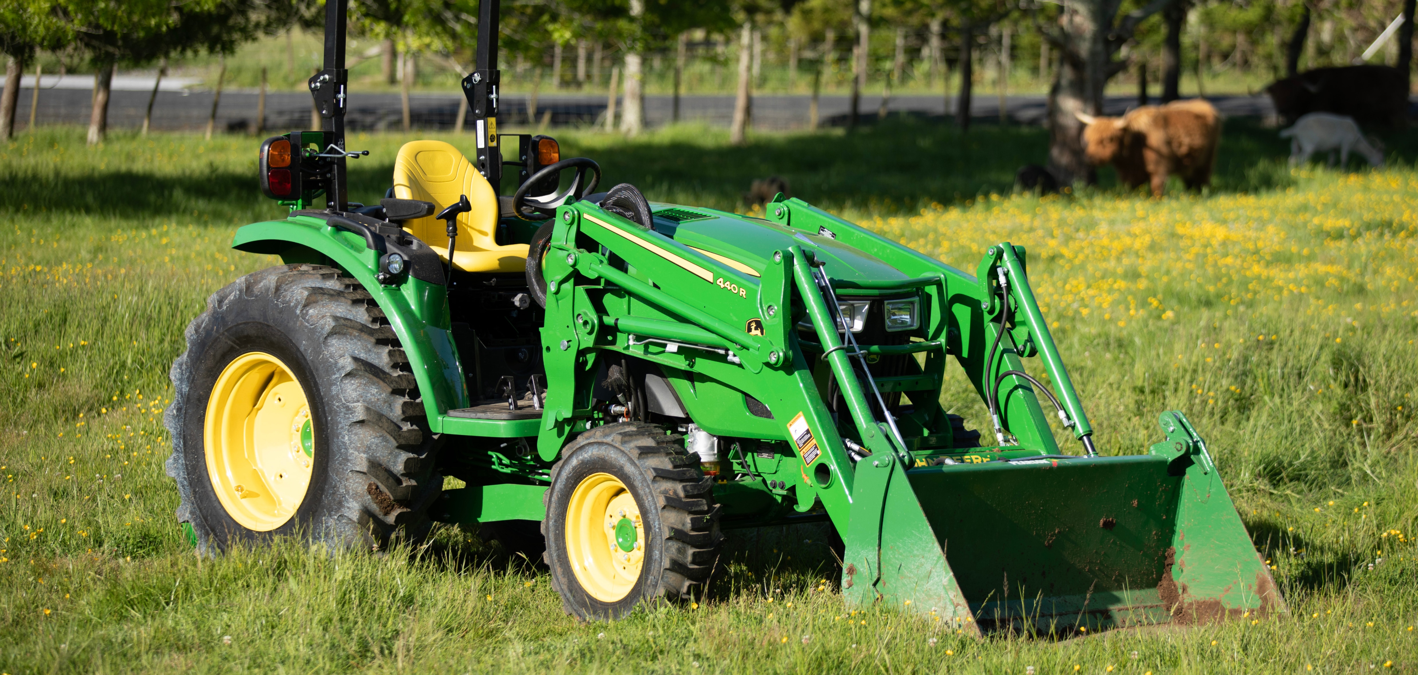 John Deere green tractor sitting on a field of grass with small yellow flowers and highland cows in the background