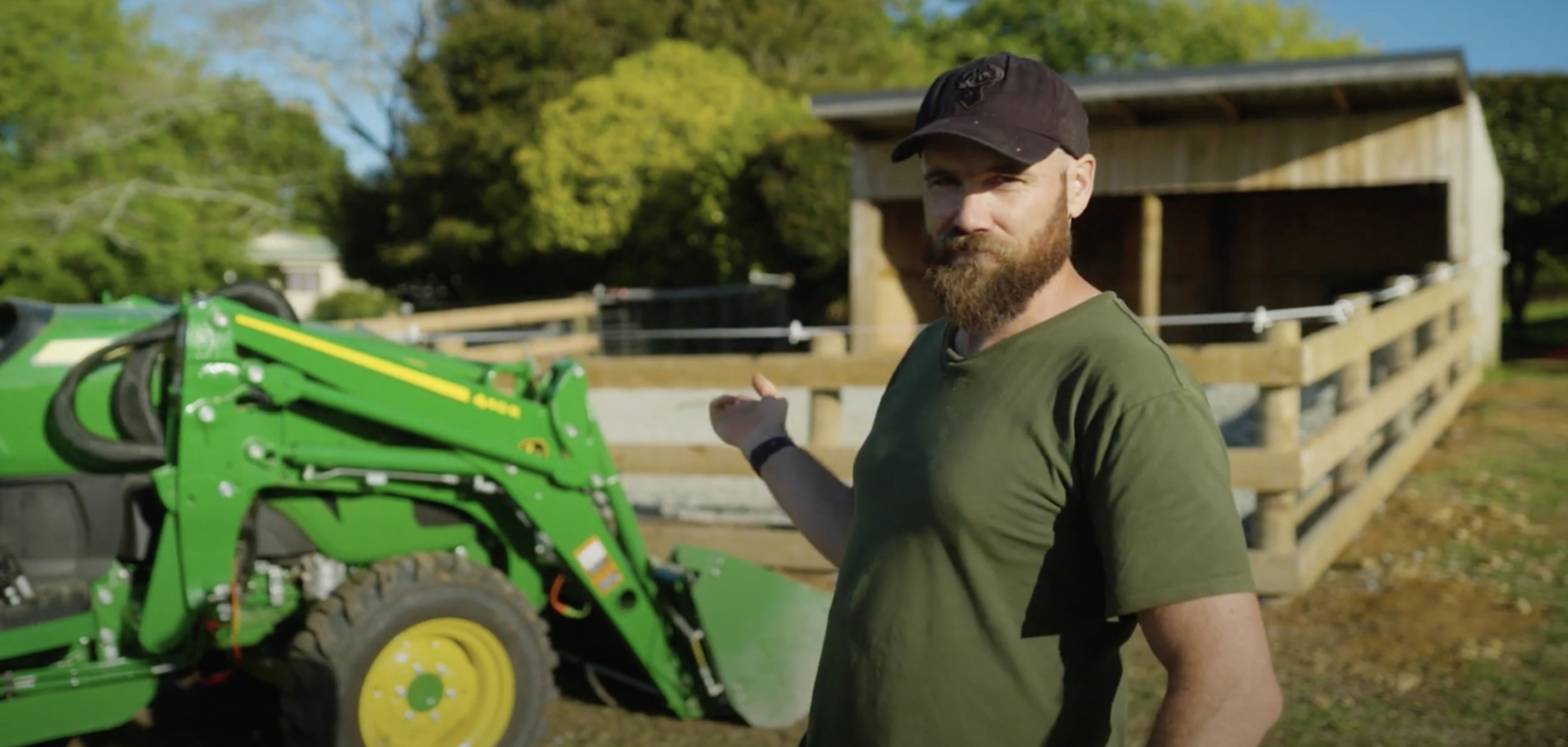 Man wearing a green shirt and black hat points towards his John Deere green tractor which is sitting in front of a fenced area and a timber shed