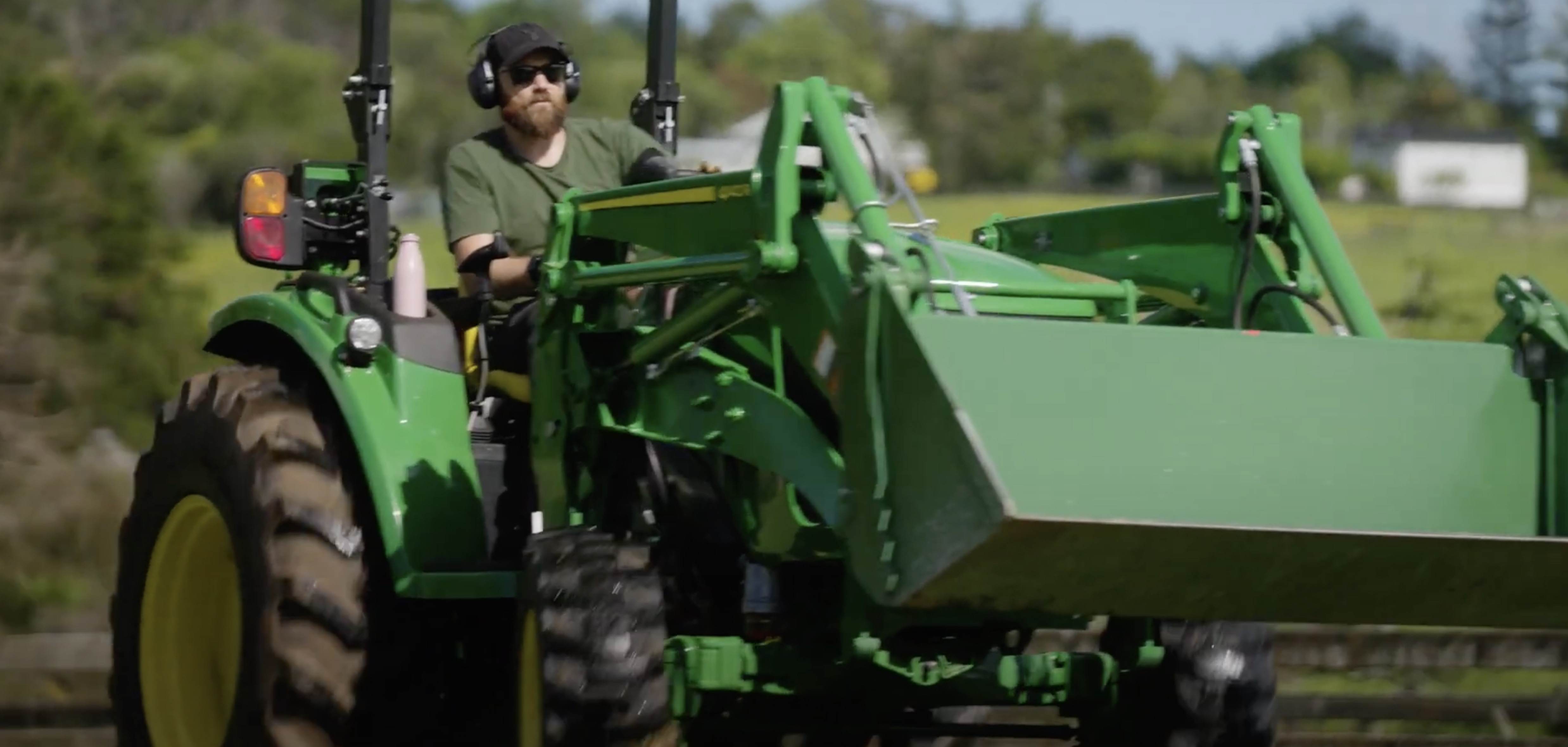 Man in green shirt, sunglasses, safety earmuffs and a black cap is driving a green John Deere tractor