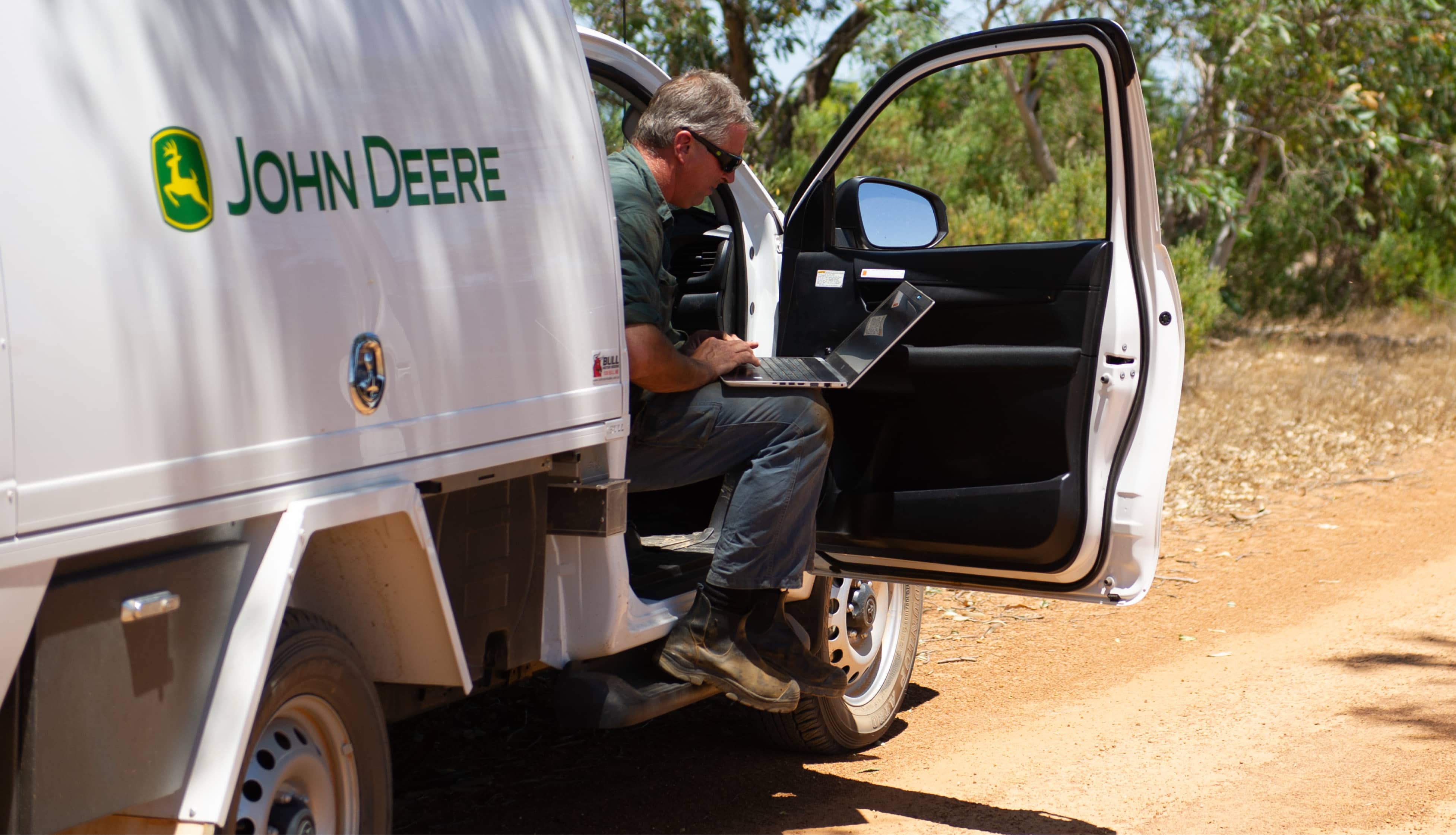 John Deere worker sits in the front of a ute with the door open into the outdoors. He is using a laptop which rests on his lap.