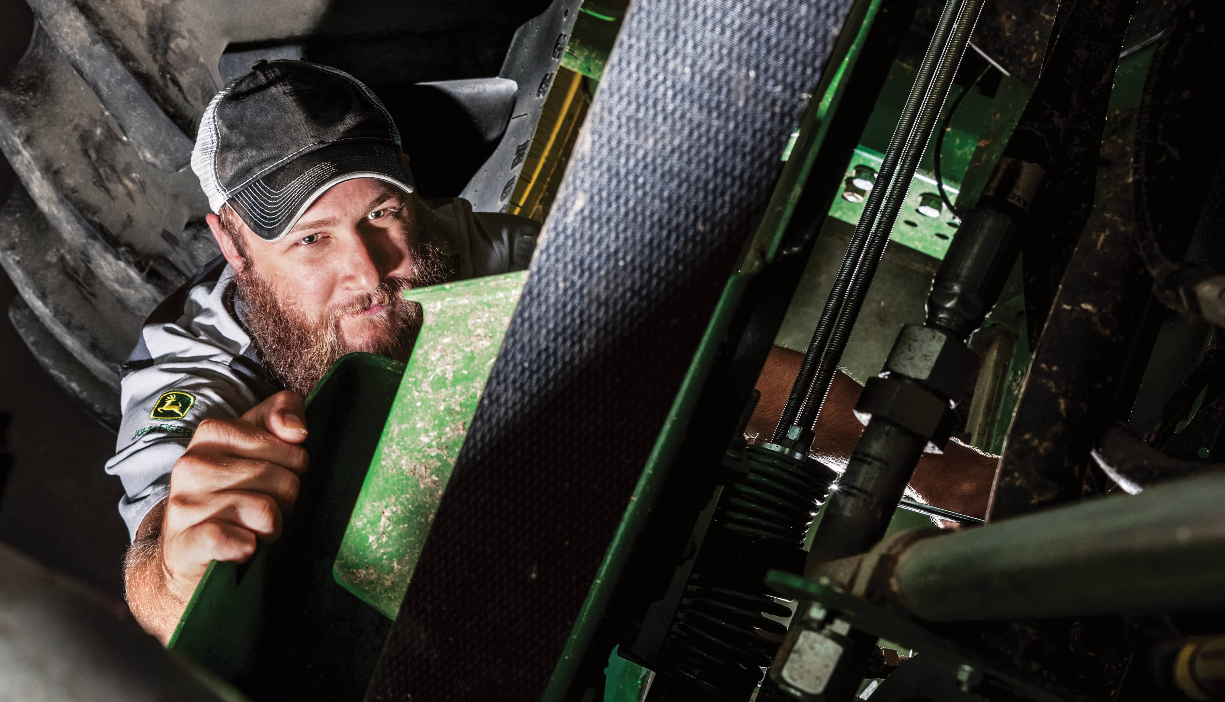 Man wearing a cap and technician uniform, underneath a tractor reaching up towards a part