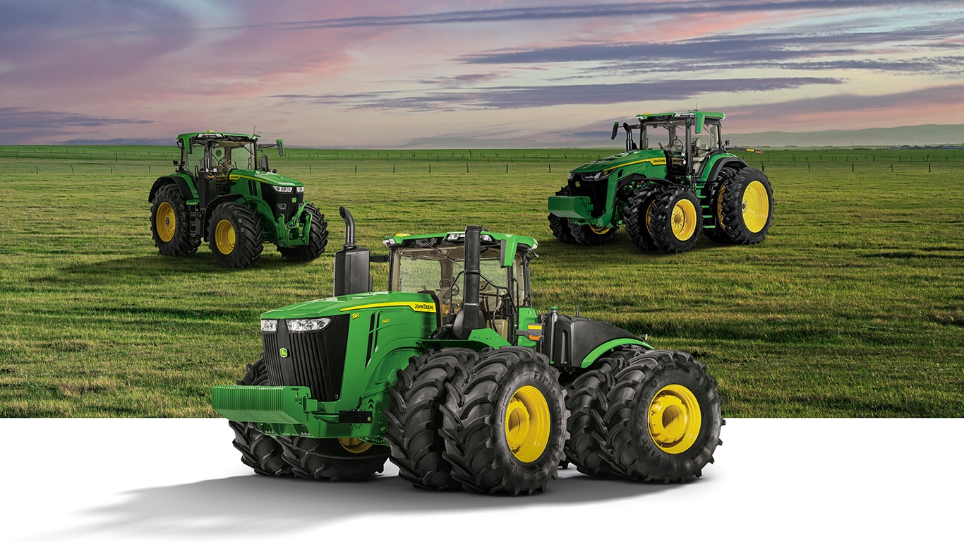 Three John Deere tractors positioned in a grassy field under a colorful sky.