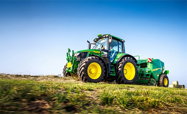 Green John Deere tractor towing a baler across a harvested field under a clear blue sky.