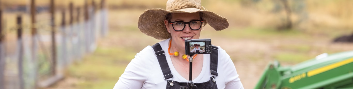 Content creator standing in front of tractor with phone attached to her overalls.