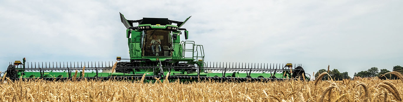 Wide photo of the front of a John Deere S7 Combine harvesting wheat with a draper head