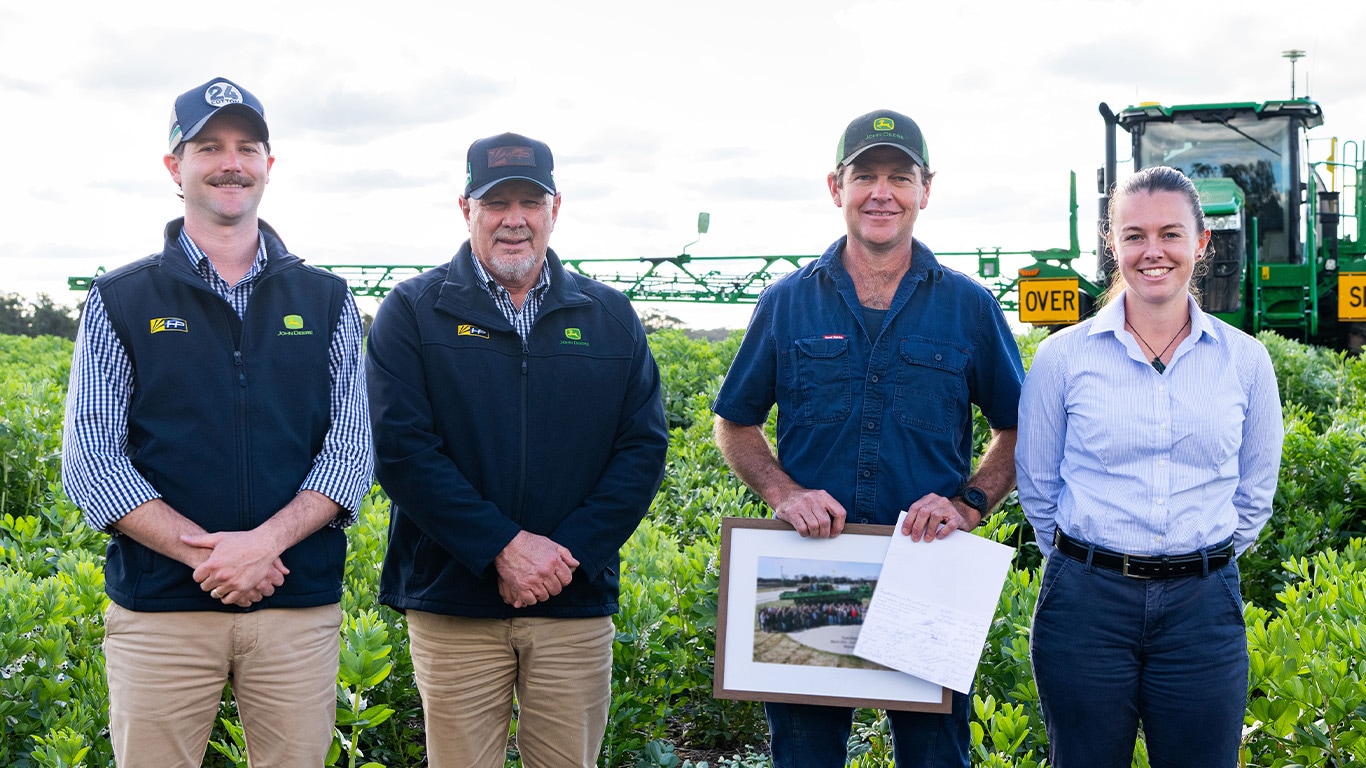 Three men and one woman standing in a field in front of a 616R sprayer, one man holds a certificate.