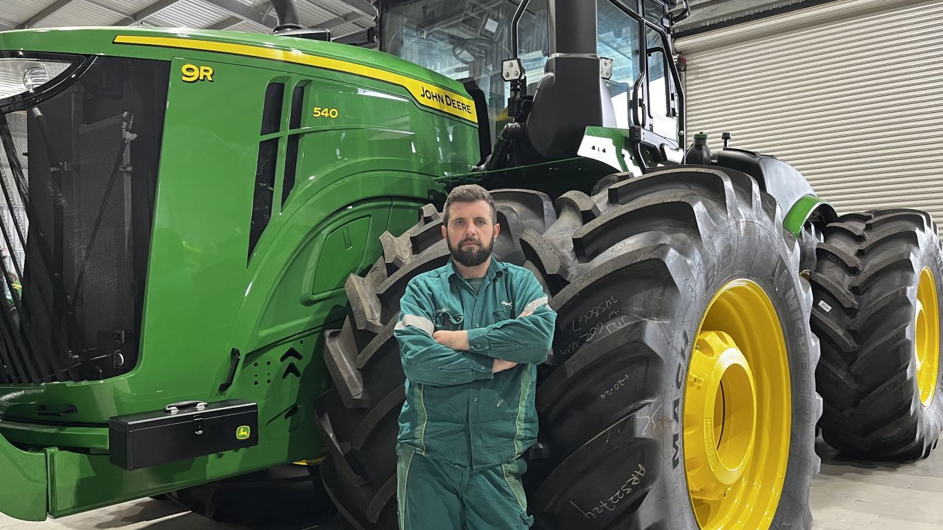 A man wearing dark green overalls standing in front of a large green tractor.