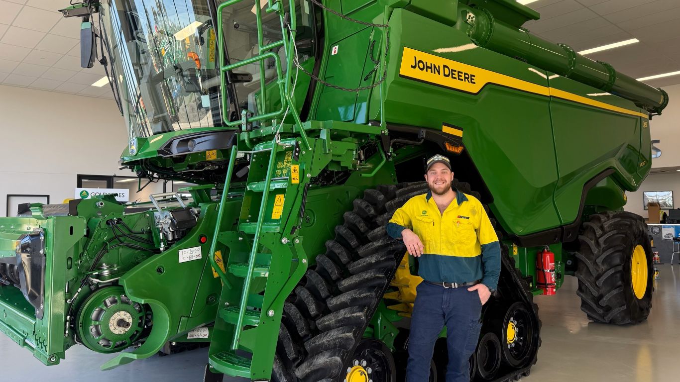 A man wearing a yellow high-visibility shirt with a cap standing in front of a large green agriculture machine.
