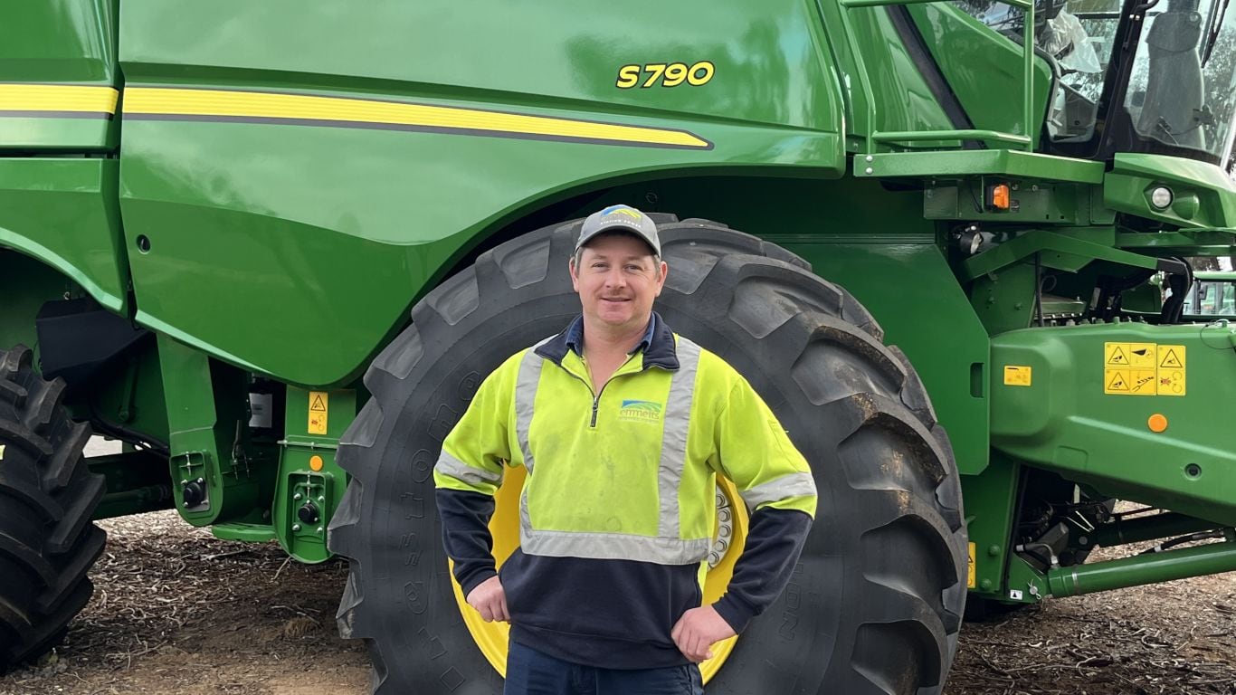A man wearing a green high-visibility jumper with a cap standing in front of a large green agriculture machine.