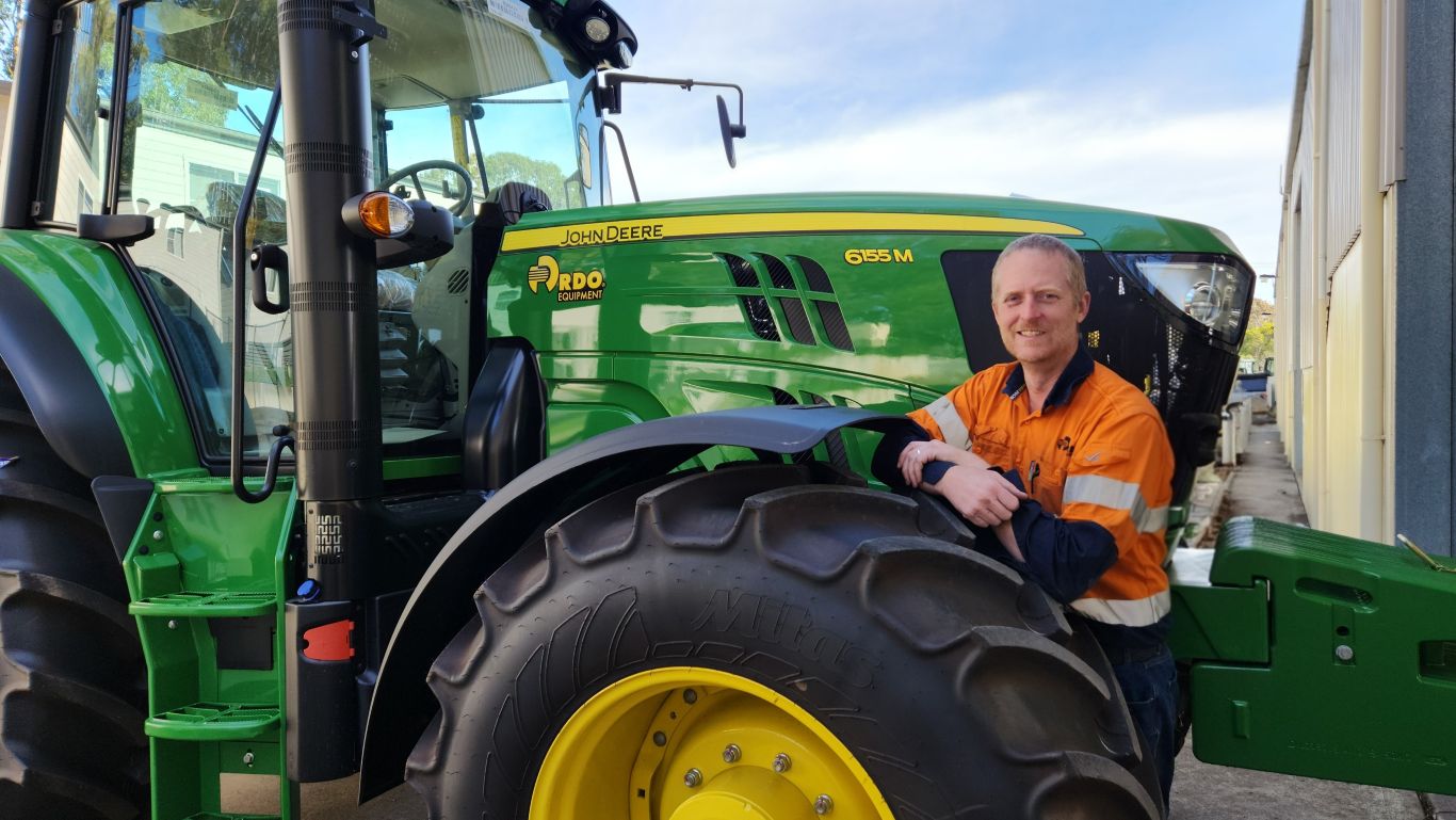 A man wearing an orange high-visibility shirt standing next to the tyre of a large green agriculture machine.