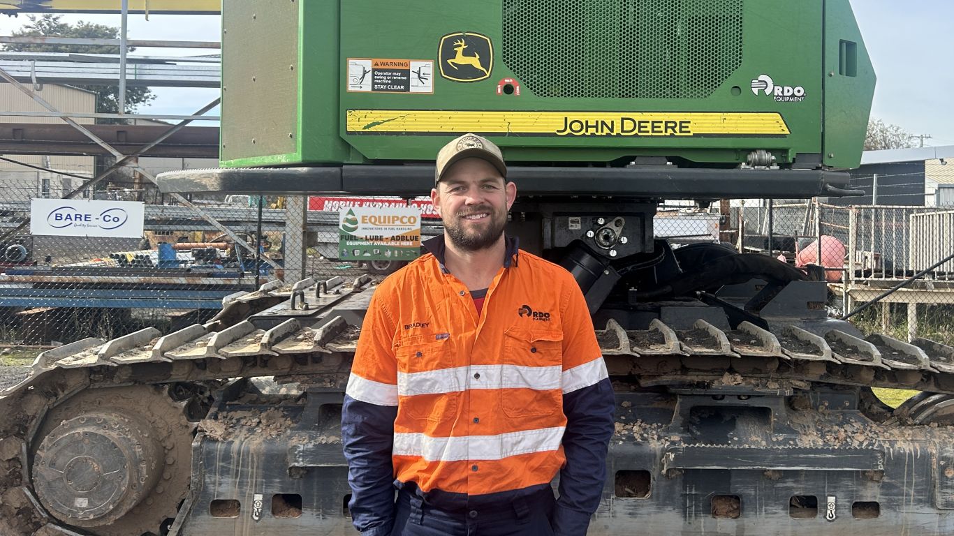 A man wearing an orange high visibility shirt standing next to a large green forestry machine.