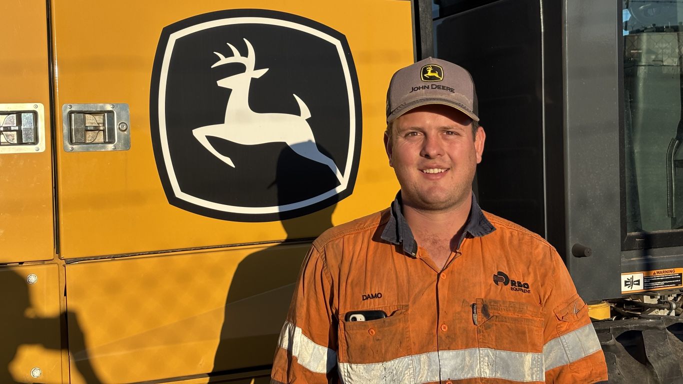 A man wearing an orange high visibility shirt standing next to a large yellow construction machine.