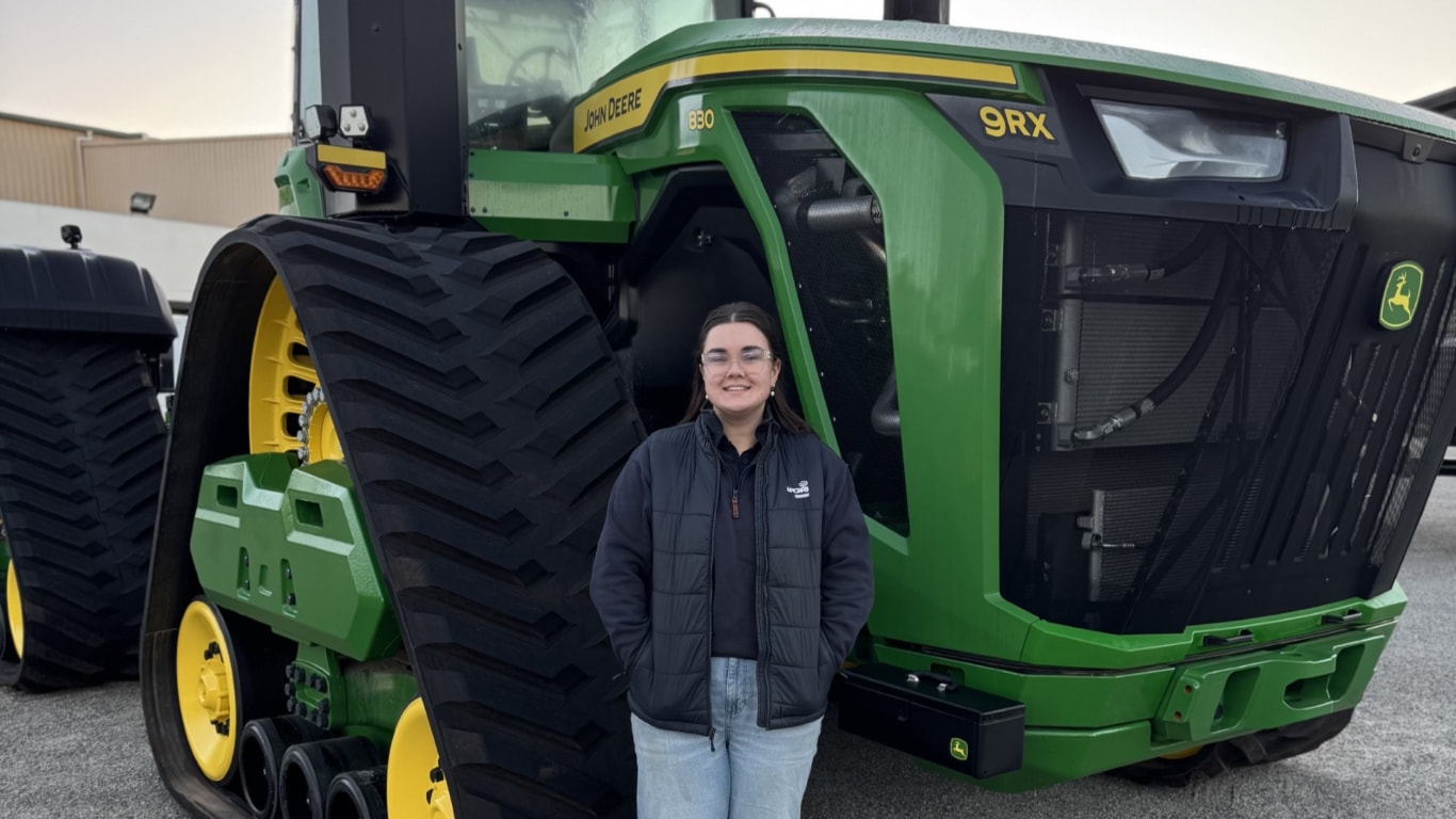 A lady wearing a black puffer jacket and blue denim jeans standing in front of a large green tractor.
