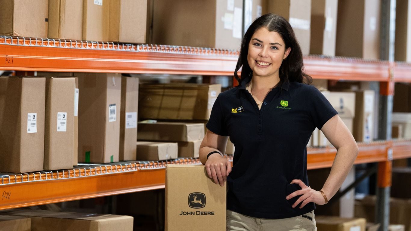A lady in a black polo shirt resting her arm on a cardboard box with orange shelves filled with cardboard boxes behind her.