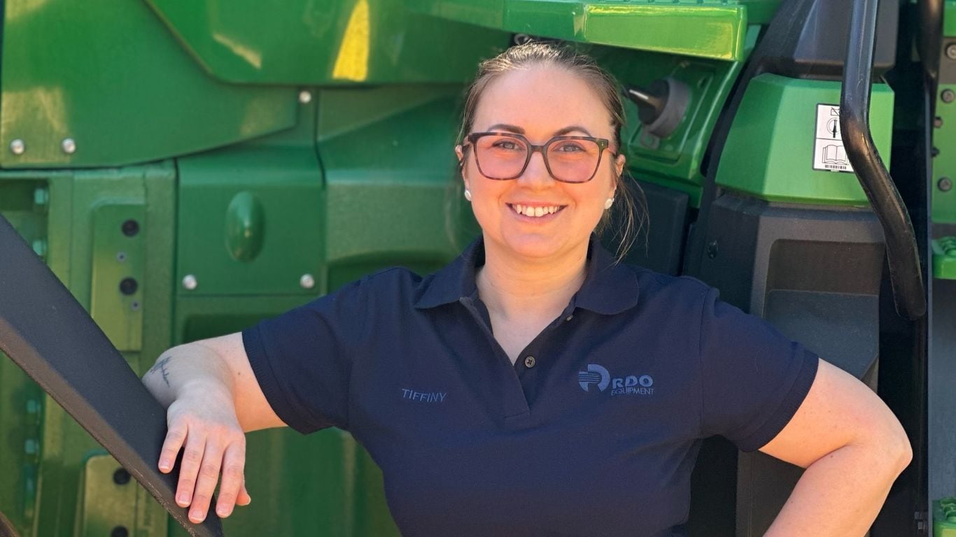 A lady in a navy-blue polo shirt leaning on green agricultural equipment.