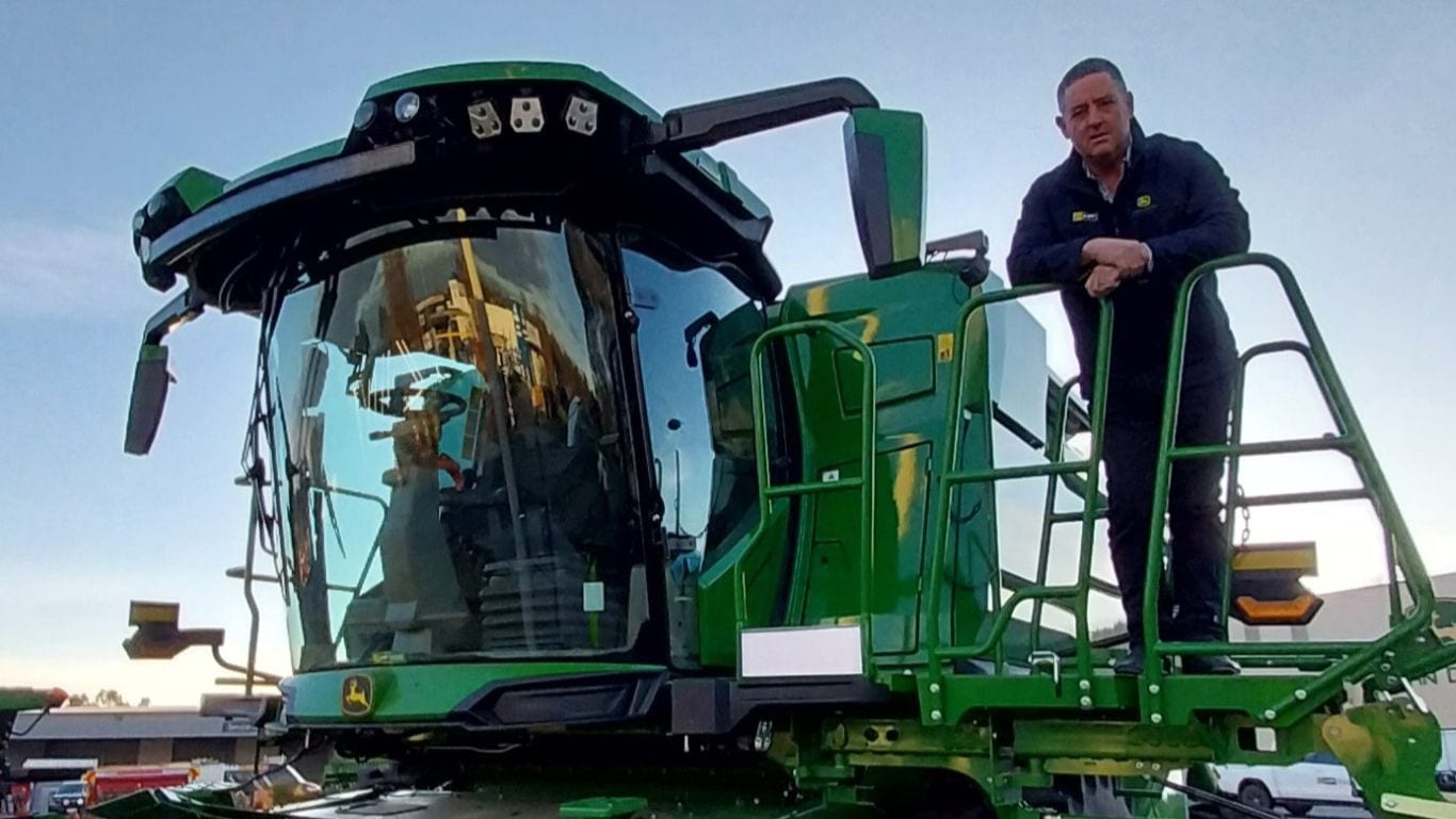 A man wearing a black jumper stands on a step near the cabin of a large green agricultural machine.