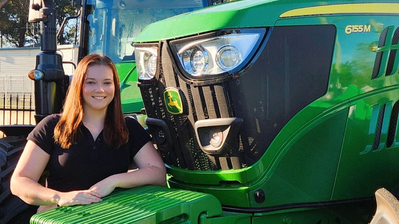 A lady wearing a black polo shirt leaning on a large green agricultural machine.