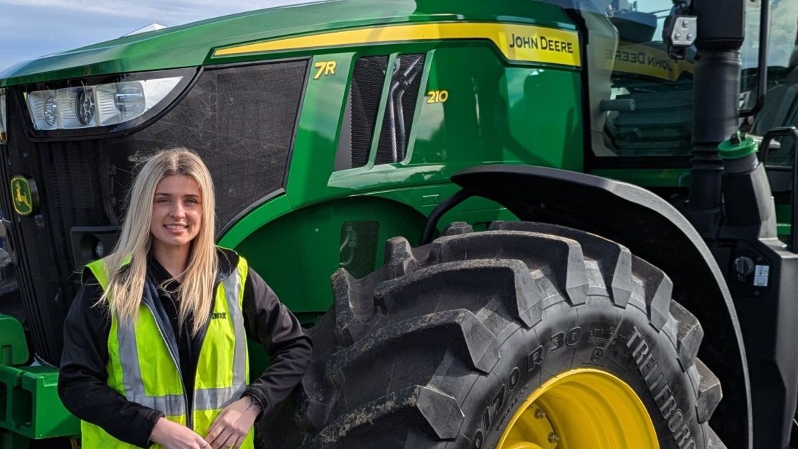 A lady wearing a yellow high visibility vest standing next to a large green agricultural machine.