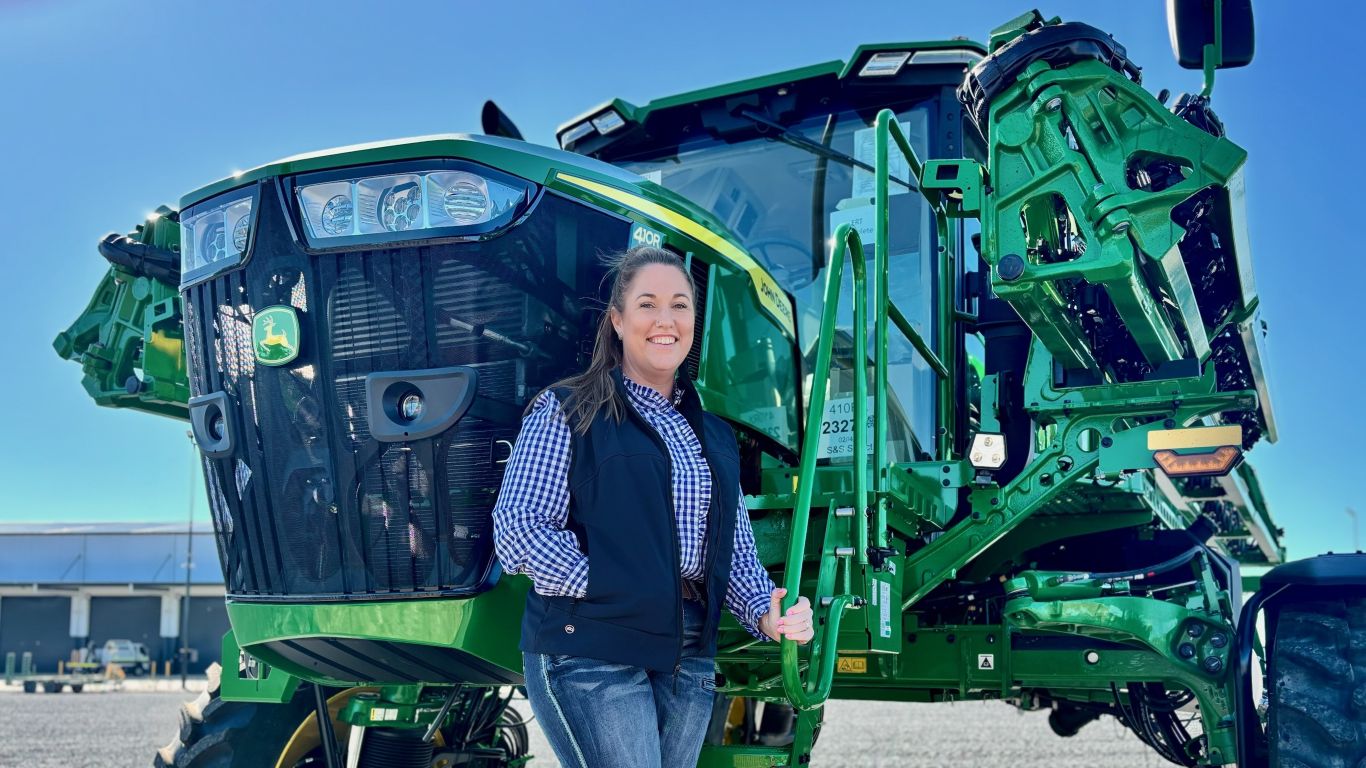 A lady with a checked shirt and blue vest standing next to a large green agricultural machine.
