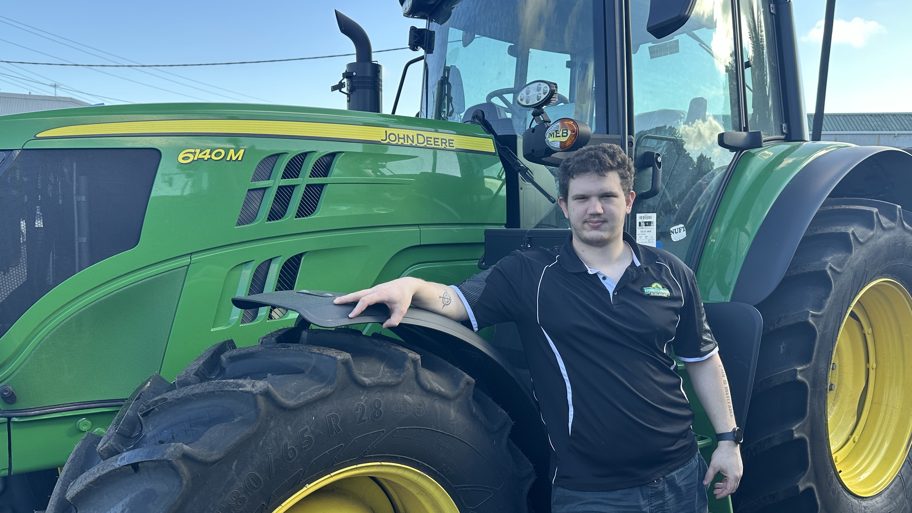 A man wearing a black polo shirt stands next to the wheel of a large green tractor.