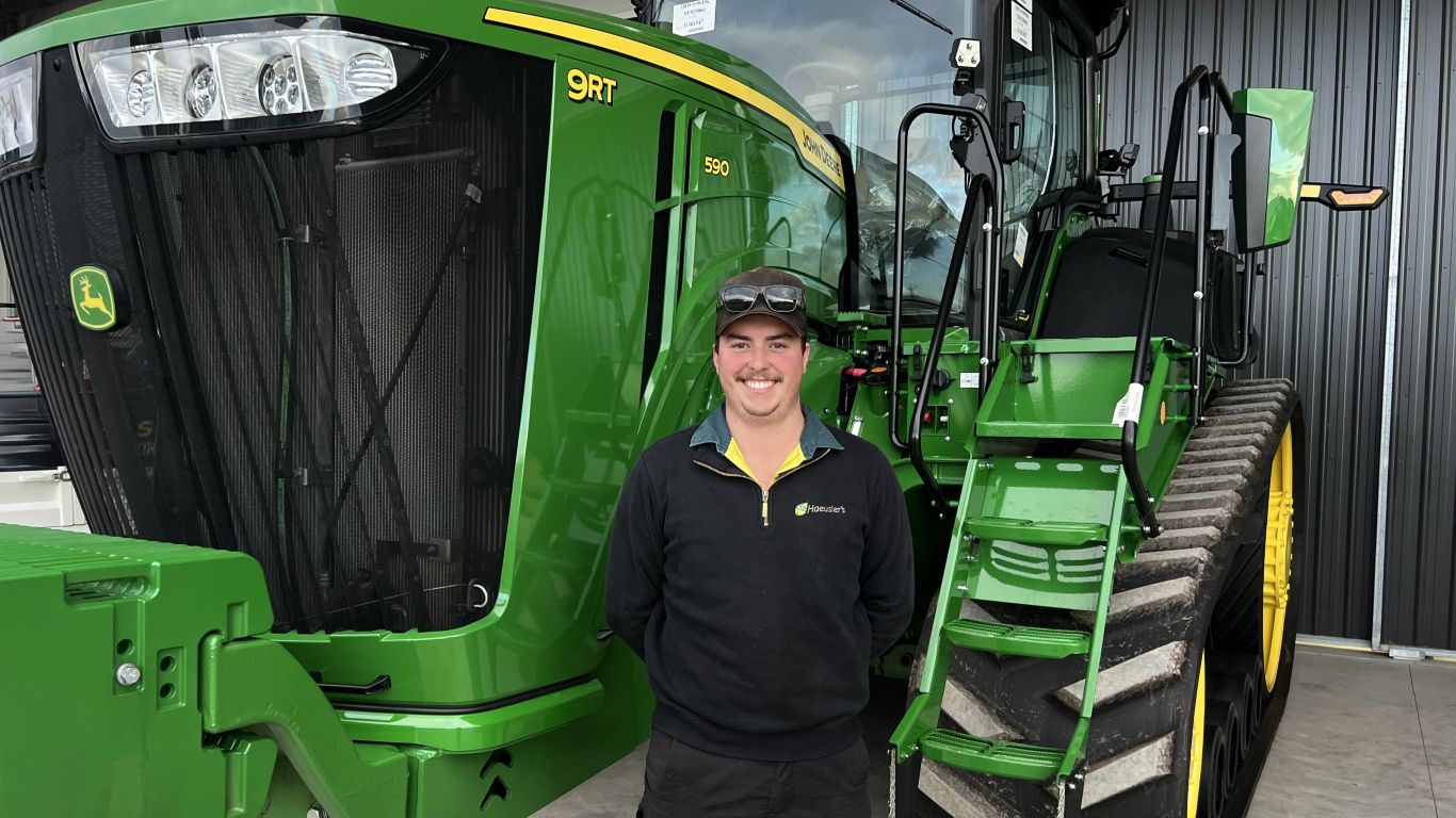 A man wearing a black jumper and a baseball cap stands next to a large green tractor.