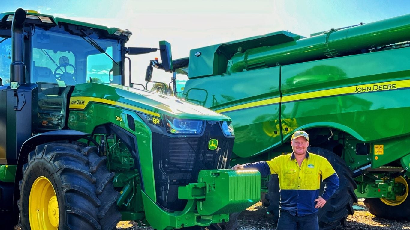 A man wearing a yellow high visibility shirt and a baseball cap, leaning on the front of a green tractor with large green agriculture machinery behind him.
