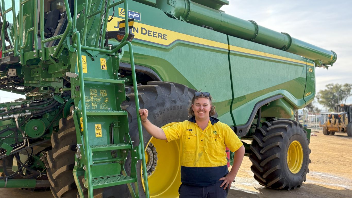 A man wearing a yellow high visibility shirt standing next to a large green agriculture machine.