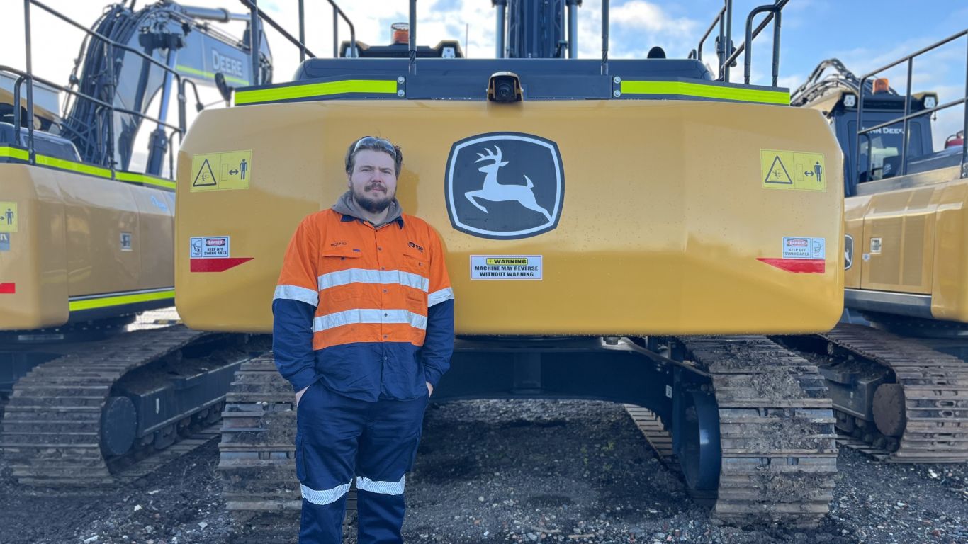 A man wearing an orange high visibility shirt standing next to a large yellow construction machine.