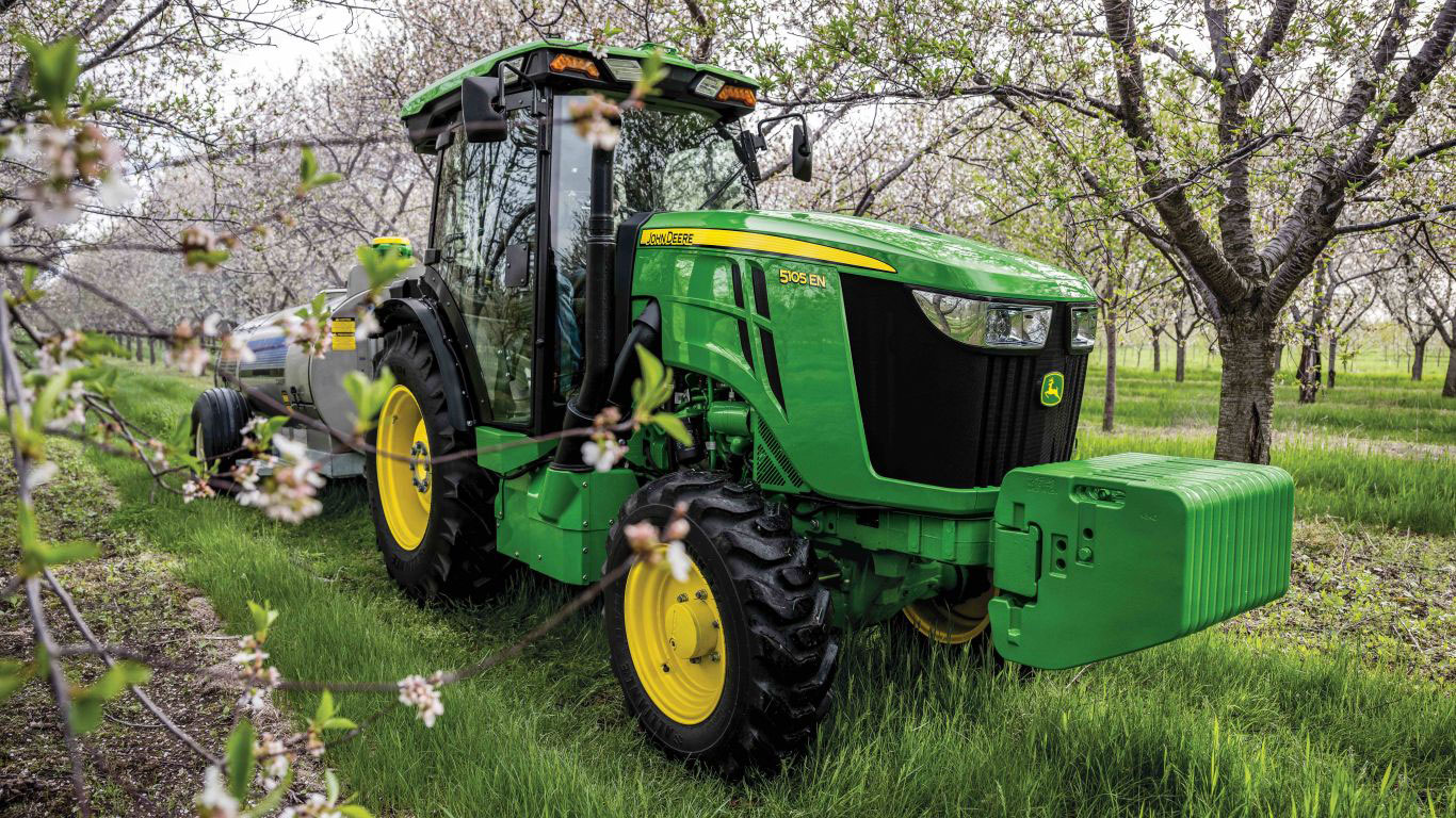 A John Deere 5105 EN tractor sits in a lush orchard, surrounded by blooming trees.