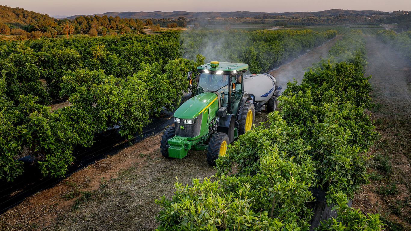 A deere tractor sprays mist among lush orange trees on a farm.