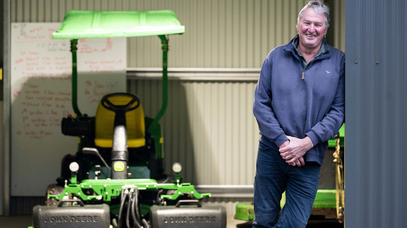 Man in navy jumper standing next to shed with John Deere equipment in the background.