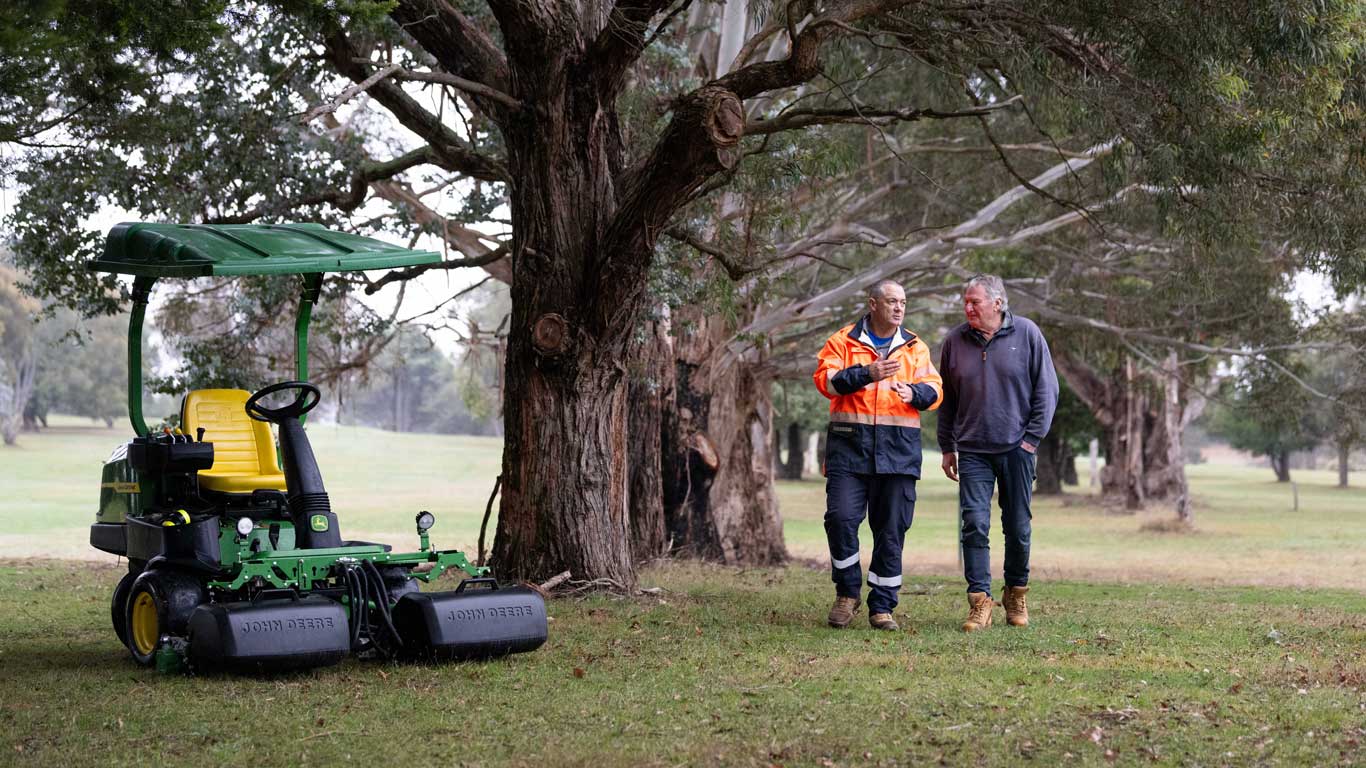 Two men, one in high vis, walking and talking on the golfing greens, with a ride on mower to the left.