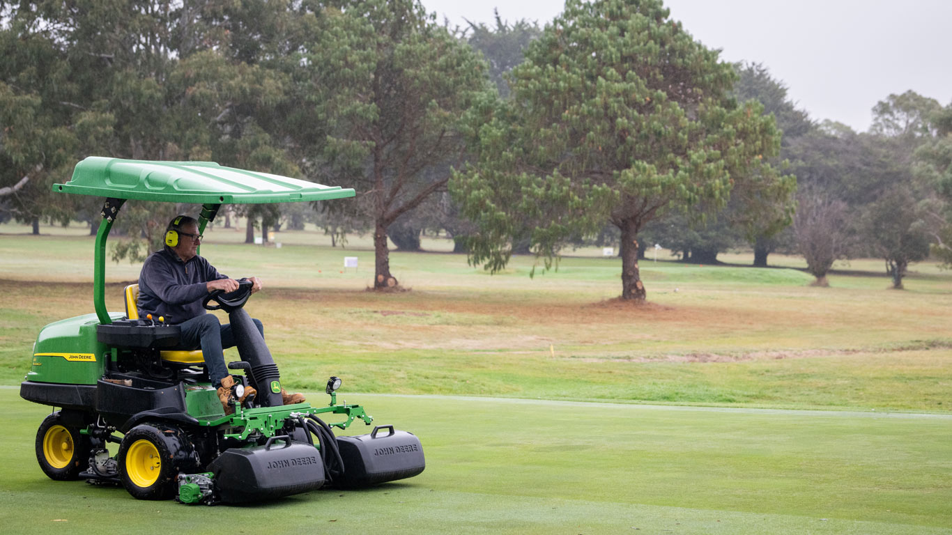 Man wearing earmuffs on a ride on mower, maintaining golfing greens.