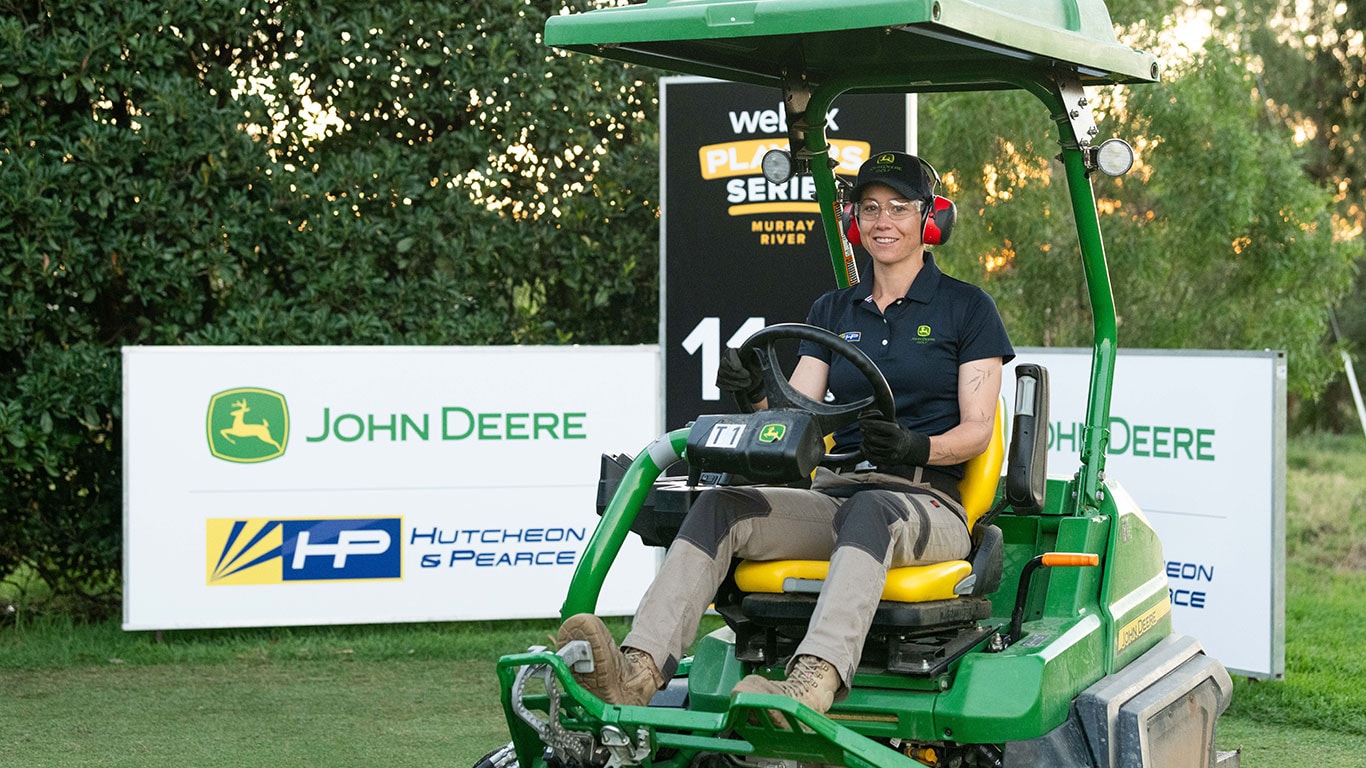 Female Volunteer raking turf in front of John Deere Signage