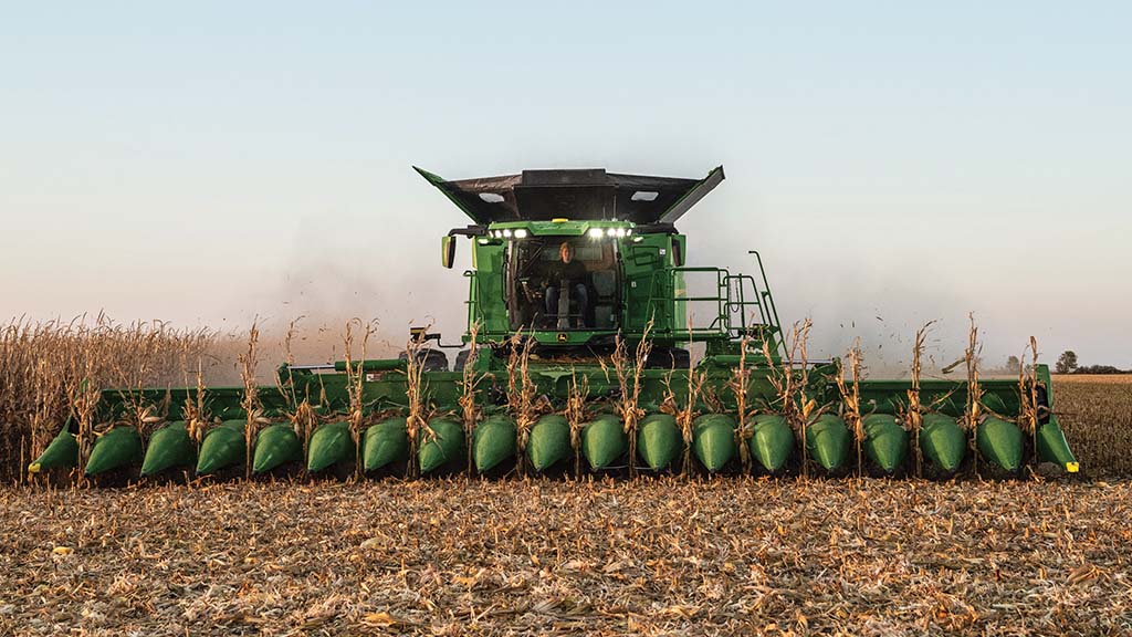 Combine harvester harvesting corn in a field, with corn stalks visible at the front, under a clear sky at dusk.