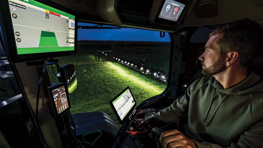 Operator monitors in-cab displays while spraying at night, with a lighted boom visible outside.